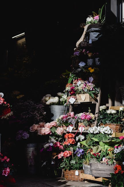 A display of fresh flowers arranged in various baskets and containers on a dark surface, with a focus on pink, white, and purple blossoms, including roses, peonies, and freesias. The flowers are arran