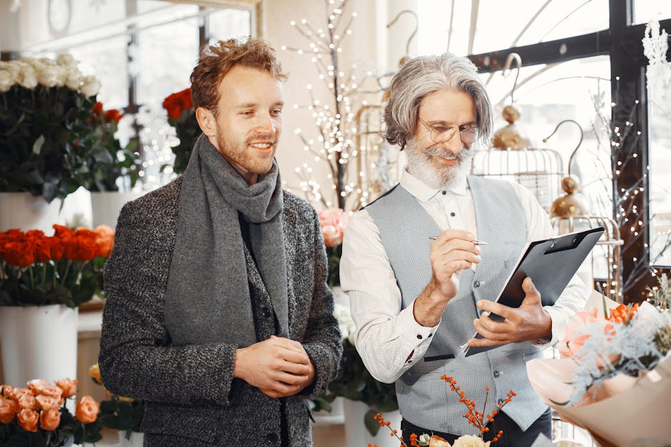 A mature male florist with long gray hair, glasses, and a beard, wearing a light blue waistcoat over a white shirt, is holding a tablet and using a stylus in a floral shop. Beside him, a younger man w