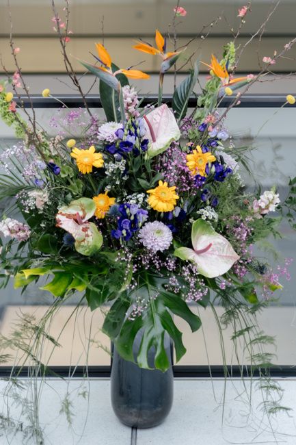A tall, vibrant floral arrangement in a dark, glossy vase featuring a variety of fresh flowers including large pink and white calla lilies, bright yellow daisies, deep blue delphiniums, and orange bir