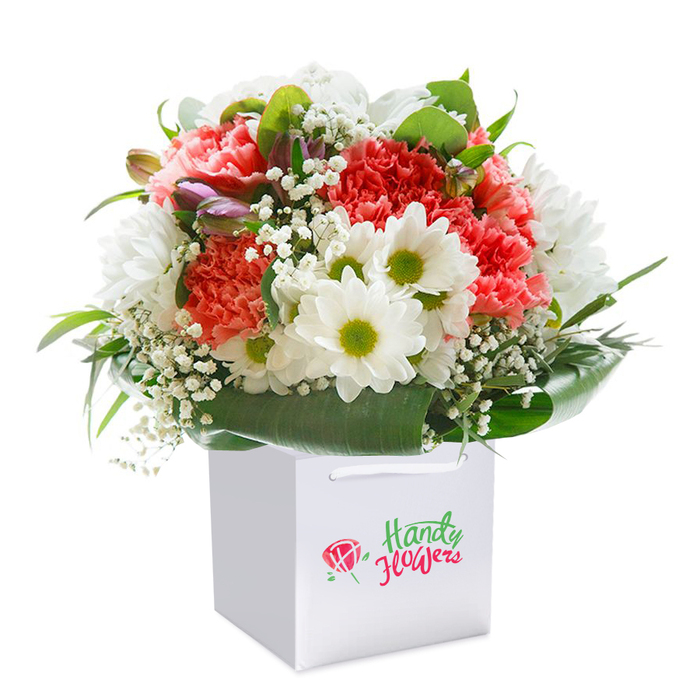 This thoughtfully arranged bouquet captures the true artistry of Shepperton floristry, presented upright in an immaculate square white gift bag and illuminated by gentle studio lighting. Central to the design are plush carnations in a palette of soft coral, peach, and bold red, each petal delicately ruffled to evoke a sense of abundant texture. Scattered between them, cheerful daisy-like chrysanthemums display their crisp white petals and lively lime-green centres-bright as a summer's stroll through Shepperton Recreation Grounds. Stems of white baby's breath weave an ephemeral tracery around the bouquet, lending a sense of airiness and natural lightness. The unique inclusion of closed buds with hints of pink and purple injects subtle depth, promising new beginnings. Surrounding it all is an elegant cradle of broad, deep green leaves, their glossy surfaces amplifying the bouquet's freshness, much like the thriving gardens that line Old Charlton Road. The sturdy, looped ribbon handles atop the gift bag make this bouquet ready for easy delivery around Shepperton, from personal milestones to heartfelt celebrations. The arrangement is captured against a pure white background, directing focus to every carefully curated detail and underscoring the exceptional quality that locals have come to expect. It's a gift that encapsulates both a sense of occasion and everyday delight, ready to enrich any Shepperton home.