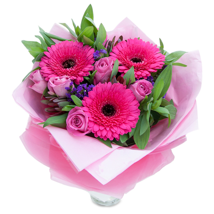 Set against a clean background, this bouquet presents three bold fuchsia Gerbera daisies at its centre - their saturated petals opening wide around dark, punchy centres that give the arrangement structure and a touch of drama. Nestled close are several roses in a softened lavender-pink or dusty-rose shade, each bloom tightly furled in a way that signals freshness and a subtle, powdery scent. Scattered through the grouping are sprigs of purple statice, their small, papery flowers lending texture and a cool contrast to the warm pinks. The foliage is abundant and green, with broad leaves that resemble salal or eucalyptus, adding a crisp, slightly dewy freshness and a calming visual frame. Thoughtful wrapping enhances the presentation: an inner layer of matte pale pink tissue provides a soft base, while an outer sheet of translucent fuchsia-pink cellophane, lightly crinkled, catches the light and adds a modern shimmer; the paper is carefully folded to cradle the stems. The lower edge reveals the rim of a clear glass vase, indicating the bouquet is prepared for instant display on a hall table or as a cheerful centrepiece. As a florist familiar with Shepperton - from quiet walks beside the river to deliveries around Shepperton Green and Laleham Road - I picture this bouquet as the perfect choice to celebrate birthdays, mark anniversaries, or simply brighten a neighbour's day, offering both visual warmth and the delicate garden-like scent of roses and fresh leaves.