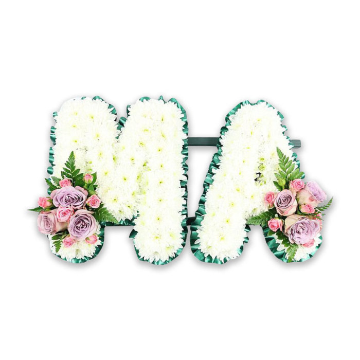 A refined MA letter tribute for a funeral or memorial service is pictured, its details clearly visible as if laid on a stand in a peaceful Shepperton chapel. Two capital letters, M and A, are formed from sturdy frames and then lavishly covered with small white chrysanthemums. These flowers are placed so closely together that they create a thick, seamless blanket of blossoms, the individual petals fanning out like tiny feathers. The chrysanthemums have a fresh, almost dewy look, with subtle shadows between each bloom that give the letters a soft yet substantial presence. Encasing both letters is a rich green border, probably pleated ribbon, carefully ruffled to produce a delicate, textured edge. The deep emerald tone of this trim cleanly outlines every angle of the M and the A, standing out against the luminous white petals and the plain background, lending the tribute an air of quiet sophistication. At the lower left side of the M and the lower right side of the A, matching clusters of accent flowers soften the design with pastel colour. There, velvety pink roses, gentle lavender roses, and petite pink spray roses are gathered in low, rounded arrangements, some blooms still tight, others open and layered. Slender fern fronds and small leaves peek through, adding a note of natural movement, reminiscent of the fresh greenery along the towpaths and quiet residential gardens around Shepperton. The composition is balanced and harmonious, suggesting both structure and tenderness. This MA tribute would be chosen by a family wishing to honour a beloved mum, mama, or someone whose initials are M.A., expressing love, gratitude, and remembrance through classic white funeral flowers gently lifted by soft pink and lilac tones.