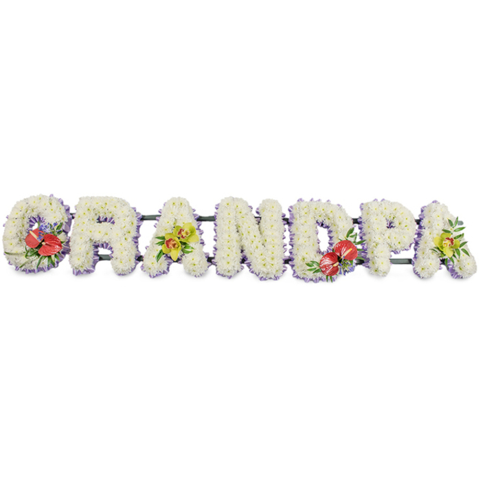 A dignified funeral tribute in this image spells the word GRANDPA in large, upright block letters, aligned in a neat horizontal row and photographed against a plain white background so all the floral work is easy to imagine. Each letter is fully covered with clusters of white chrysanthemums, the small blooms pressed closely together to form a continuous, cushion-like texture, with their rounded petals softening the strong shapes of the lettering. The white flowers glow gently against the neutral background, suggesting peace, innocence, and a quiet farewell to a cherished grandfather. At the base of each letter, a fine band of pale lavender ribbon wraps around the frame, peeking out just enough to create a slender lilac border that defines the contours and adds a subtle, soothing note of colour. Behind the letters, dark green supporting bars run from one end of the word to the other, giving the tribute a sense of stability and making it easy to picture the arrangement resting on trestles at a church or crematorium service in Shepperton. To relieve the expanse of white, the florist has introduced thoughtfully placed accent sprays: vibrant red anthuriums decorate the G and the D, their glossy, heart-shaped spathes and central spadices framed by rich greenery, forming focal points of passion and remembrance. On the first A and final A, clusters of yellow-green orchids sit lightly among the chrysanthemums, their distinctive blossoms and lime-tinged petals lending elegance and a feeling of ongoing life. The overall design is both strong and tender, a personalised floral name tribute that might be chosen by a Shepperton family to stand by the coffin or at the front of the service, quietly honouring a granddad whose influence was steady, loving, and deeply felt.